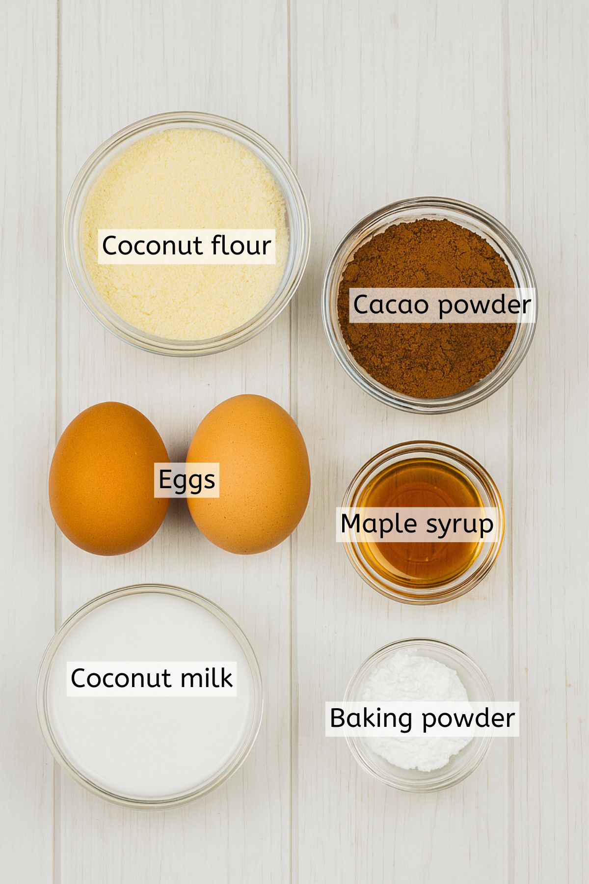 Coconut flour. cacao, maple syrup, and milk in bowls with eggs beside them and text labels over everything.