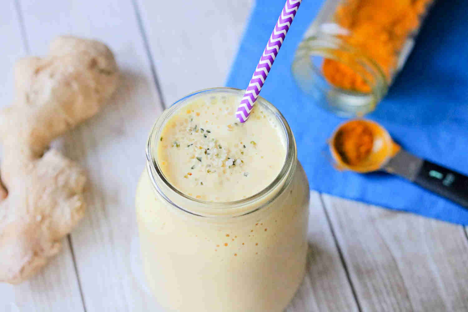 turmeric peach smoothie in a mason jar with a purple straw. Turmeric root and powder and ginger root around it