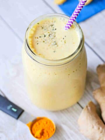 turmeric peach smoothie in a mason jar with a purple straw. Turmeric root and powder and ginger root around it