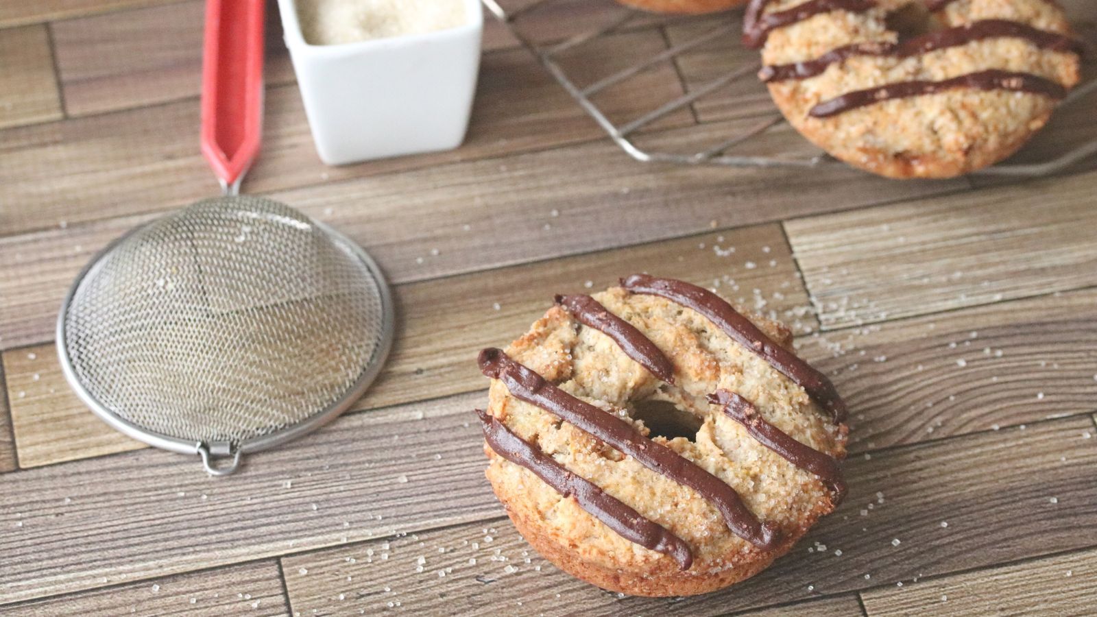 banana protein donuts topped with chocolate. One in the middle with a strainer and sugar around it the others on a cooling tray