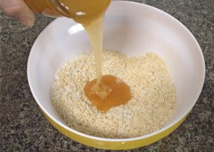 Honey being poured into a yellow mixing bowl with protein powder and coconut flakes