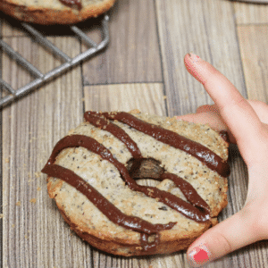 Banana protein donut iced with chocolate sauce with a toddlers hand grabbing it and more donuts on a cooling rack behind them