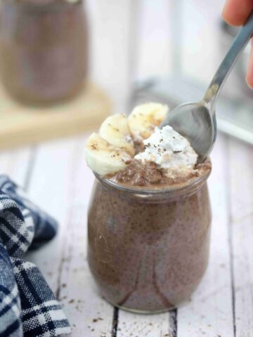 Chocolate chia pudding in a jar with whole30 whipped cream and bananas on top and a spoon going into the jar.