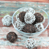 Chocolate coconut energy balls with dates in a glass bowl with three energy balls beside the bowl and coconut around it