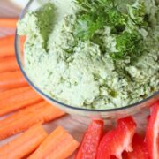 Green chickpea hummus in a glass bowl with parsley on top and carrots, red pepper and celery around it.