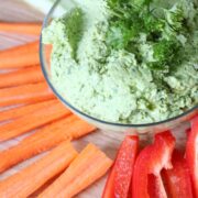 Green chickpea hummus in a glass bowl with parsley on top and carrots, red pepper and celery around it.