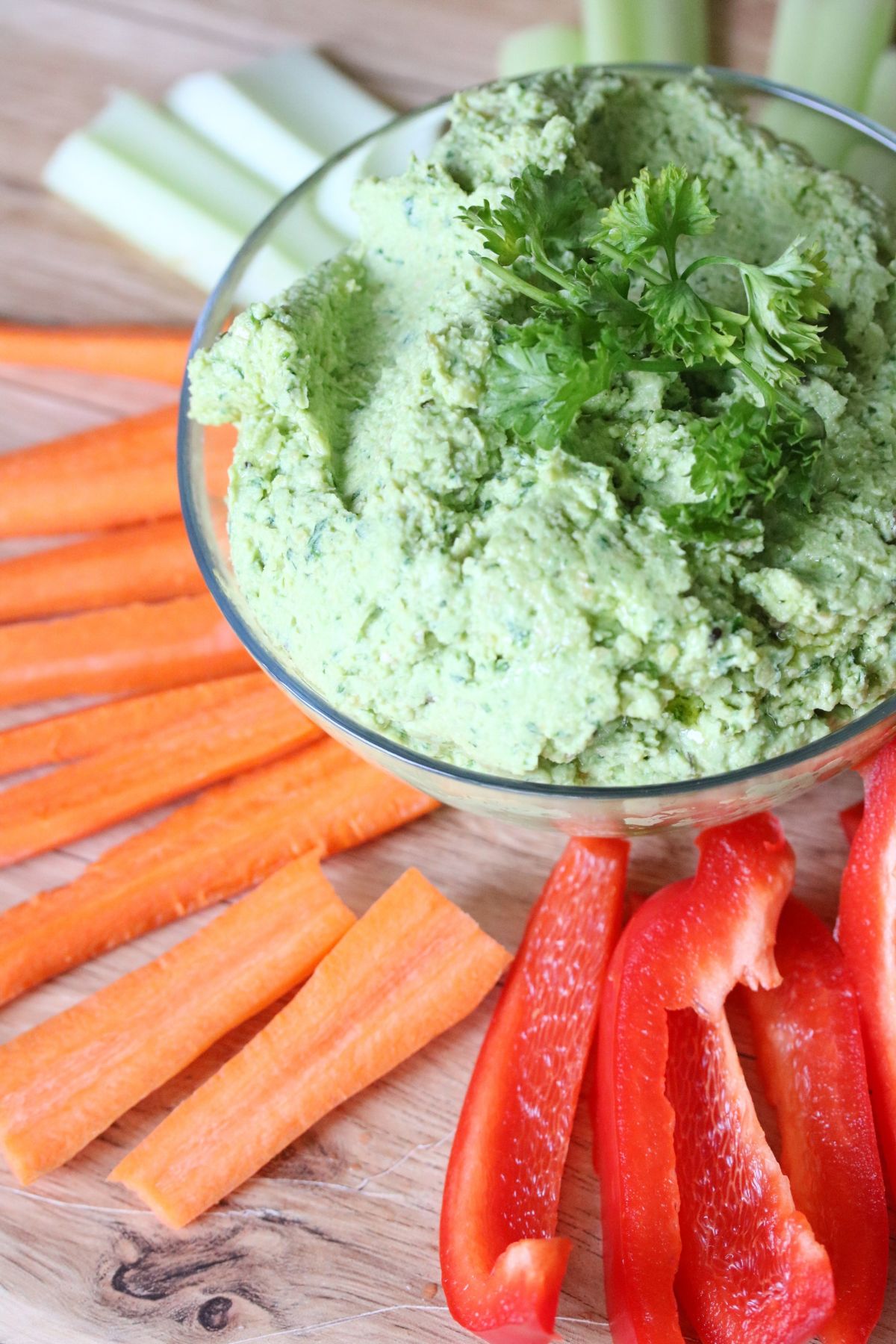 Green chickpea hummus in a glass bowl with parsley on top and carrots, red pepper and celery around it.