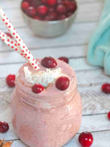 Apple cranberry smoothie i a mason jar with whole30 whipped cream and cranberries on top and cranberries around the jar and straws in it.