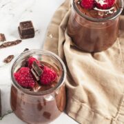 Two mason jars filled with chocolate pudding on a tan linen and topped with rapsberries and berries around them.