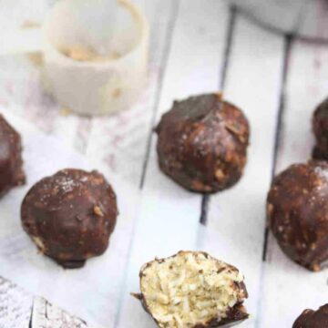 Multiple chocolate protein coconut balls lined up and one with a bite out of it, with protein powder off to the side.