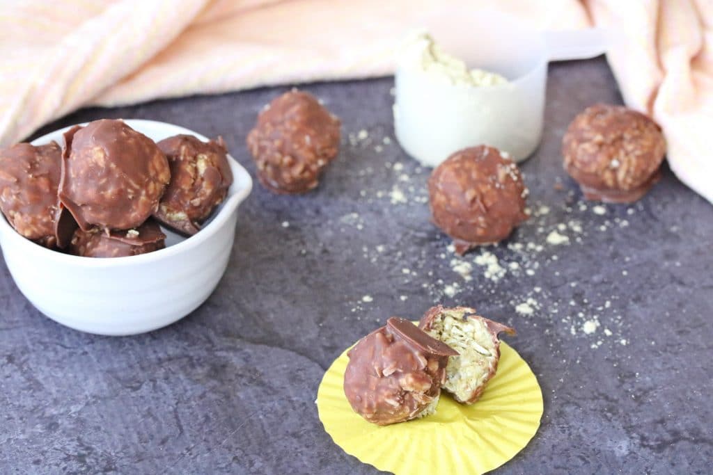 No-bake chocolate covered protein ball on a yellow cupcake cup with more balls in a bowl behind and protein powder