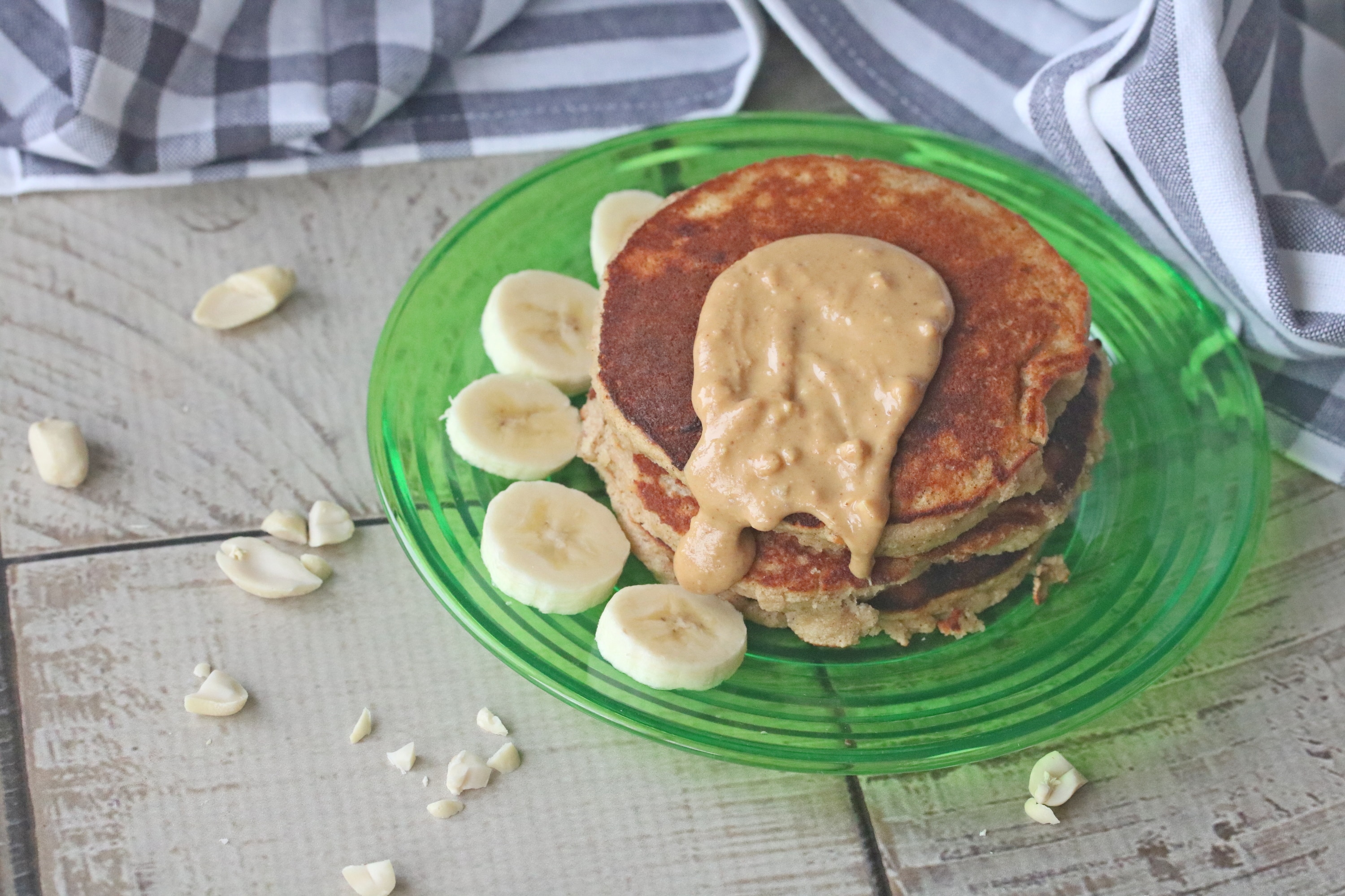 Three peanut butter banana pancakes stacked on a green plate with peanut butter on top and sliced bananas on the plate with a blue linen behind the plate.