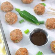 Multiple turkey meatballs on parchment paper with a jalapeno, steel tin of sauce, toothpicks, and green onions around them.