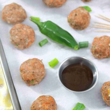 Multiple turkey meatballs on parchment paper with a jalapeno, steel tin of sauce, toothpicks, and green onions around them.