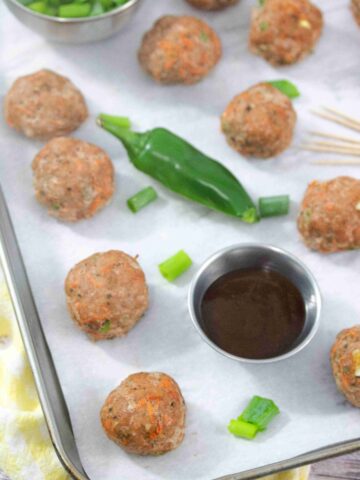 Multiple turkey meatballs on parchment paper with a jalapeno, steel tin of sauce, toothpicks, and green onions around them.