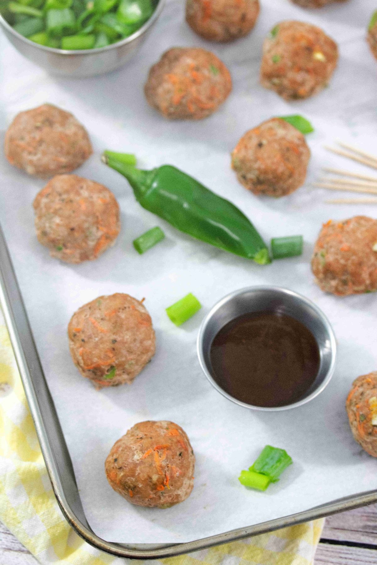 Multiple turkey meatballs on parchment paper with a jalapeno, steel tin of sauce, toothpicks, and green onions around them.