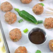 Multiple turkey meatballs on parchment paper with a jalapeno, steel tin of sauce, toothpicks, and green onions around them.