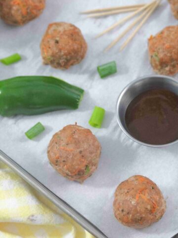 Multiple turkey meatballs on parchment paper with a jalapeno, steel tin of sauce, toothpicks, and green onions around them.