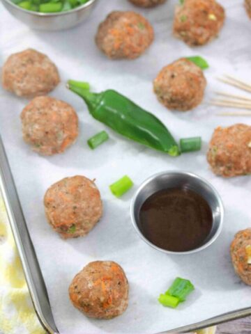 Multiple turkey meatballs on parchment paper with a jalapeno, steel tin of sauce, toothpicks, and green onions around them.