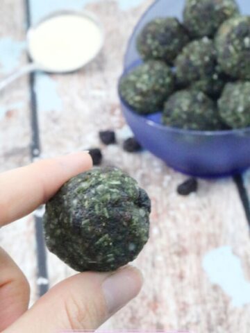 Two fingers holding up a superfood energy ball with more balls in a blue bowl behind them