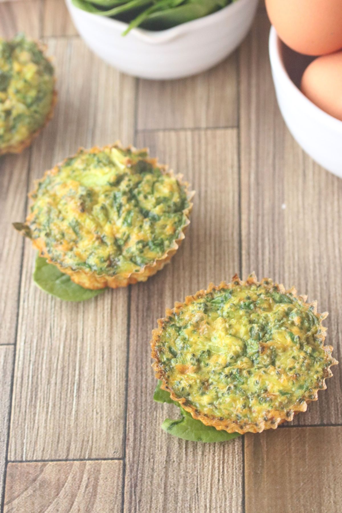 Dairy-free egg muffins in a line seated on spinach with eggs and spinach in white bowls.