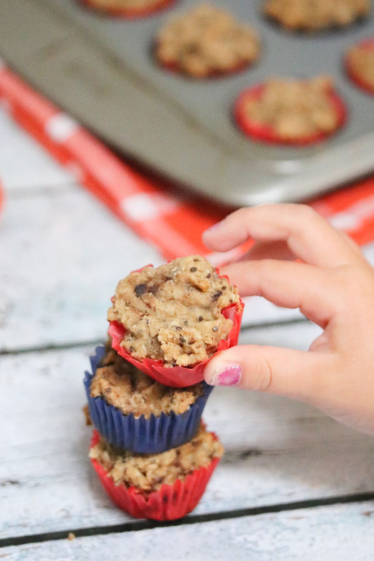 Three bite-sized toddler muffins stacked on top of each other with a toddlers hand reaching for them andwith chia seeds and a white ramekin with more muffins behind them.