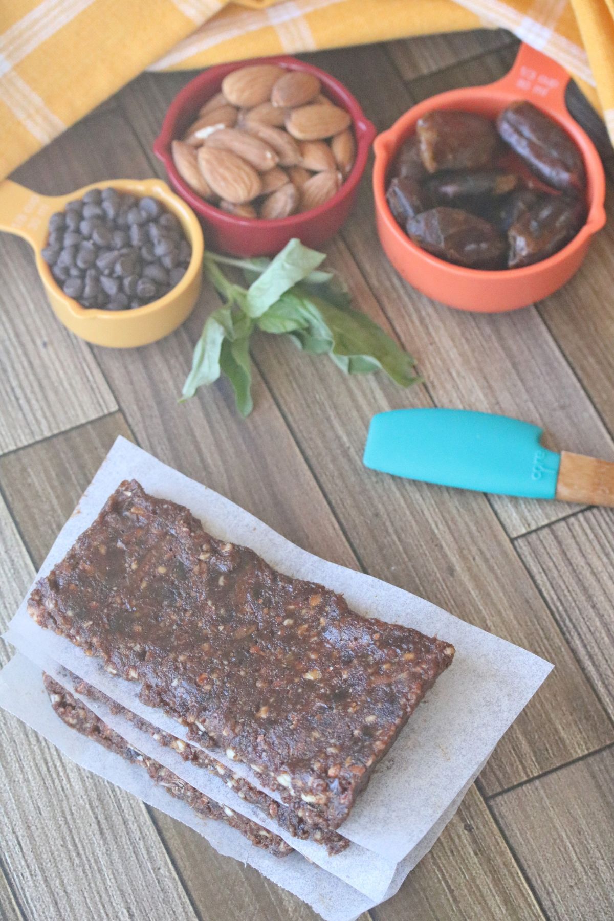 Three chocolate energy bars stacked with parchment paper between them and ingredients in measuring cups behind them.