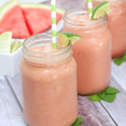 Three mason jars filled with watermelon smoothie and topped with green straws and lime sitting on mint leaves and fresh watermelon behind them