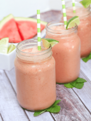 Three mason jars filled with watermelon smoothie and topped with green straws and lime sitting on mint leaves and fresh watermelon behind them