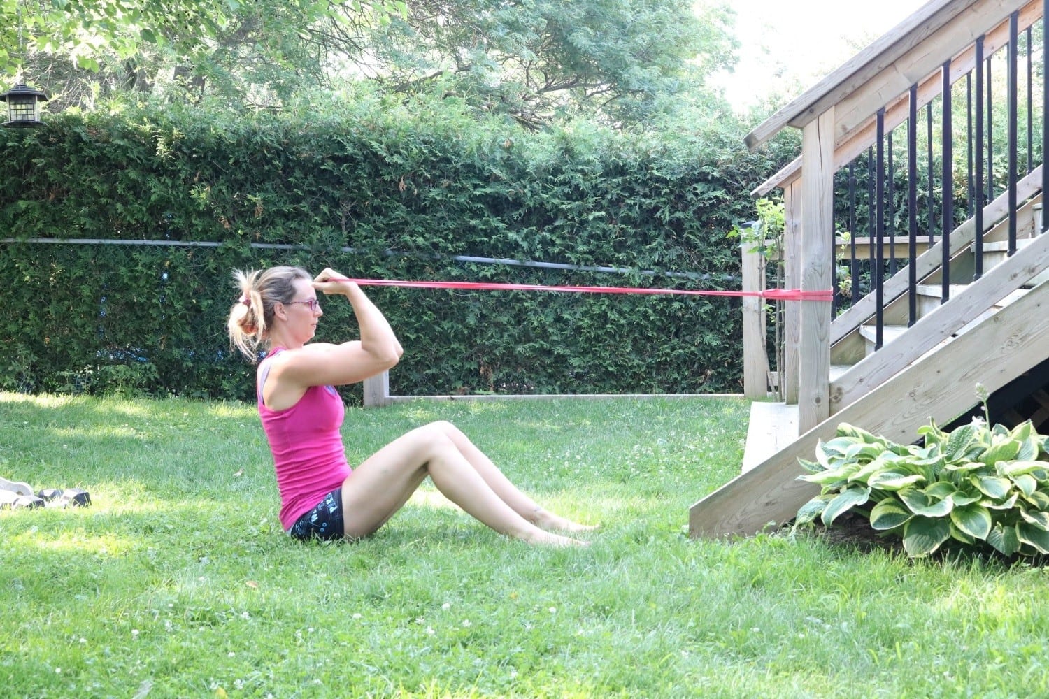 women in pink shirt and black shorts sitting on grass doing a bicep curl with a red resistance band