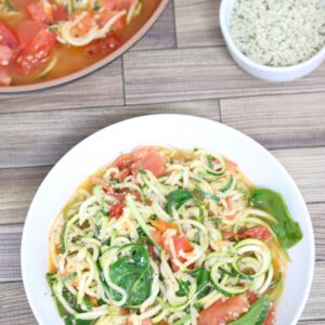 Zucchini and tomato pasta with spinach in a white bowl with more in a pan behind it