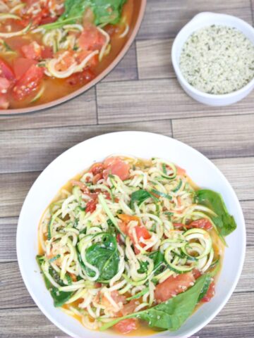 Zucchini and tomato pasta with spinach in a white bowl with more in a pan behind it