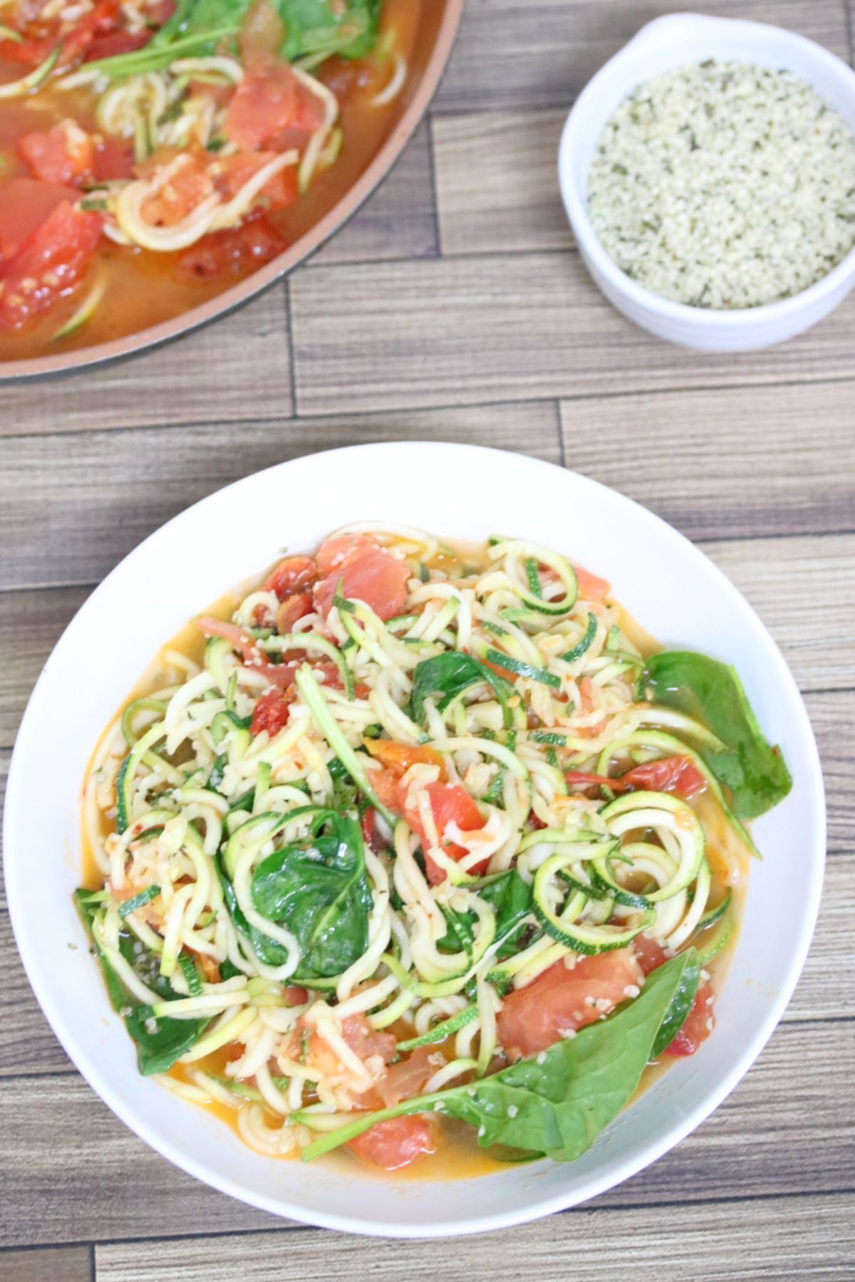 Zucchini and tomato pasta with spinach in a white bowl with more in a pan behind it
