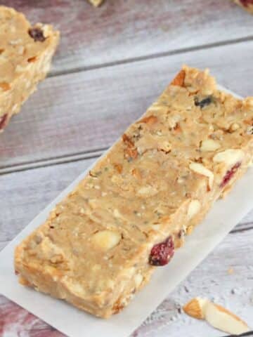 One homemade protein bar on parchment paper with more protein bars, cranberries and protein powder tub around it.
