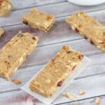 One homemade protein bar on parchment paper with more protein bars, cranberries and protein powder tub around it.