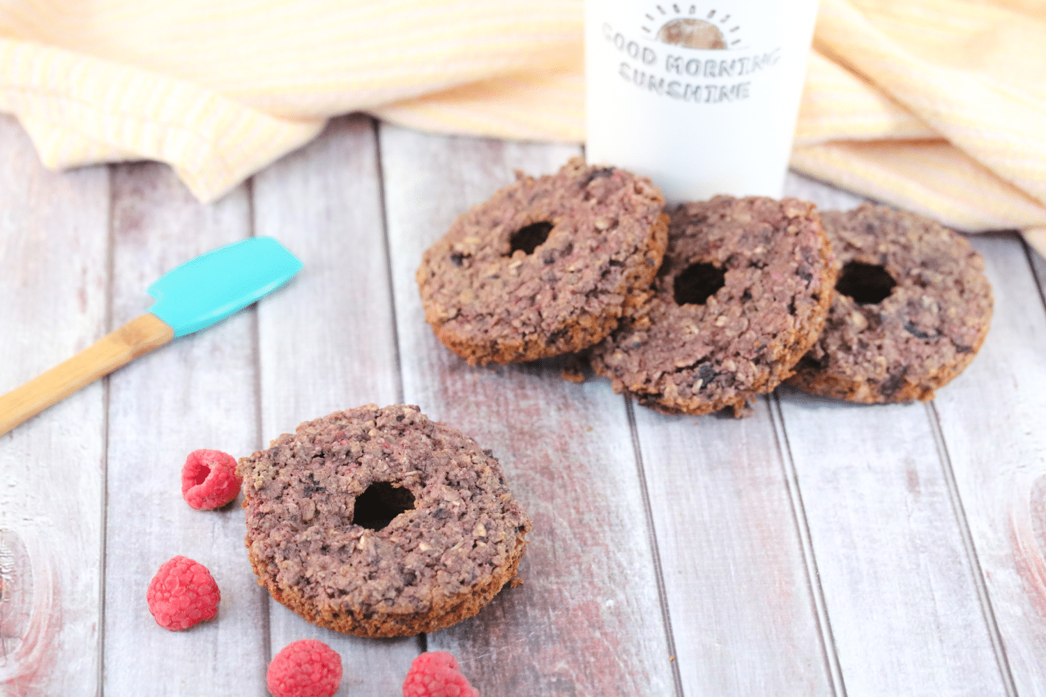 One gluten-free donut with raspberries around i t and three more around a coffee mug with an orange linen