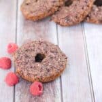 One gluten-free donut with raspberries around i t and three more around a coffee mug with an orange linen.