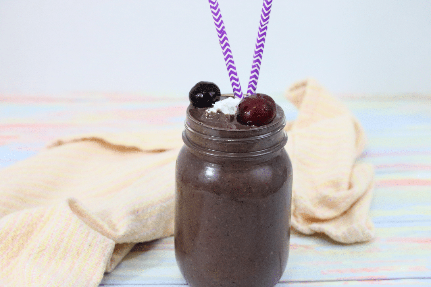 chocolate cherry smoothie in a mason jar topped with coconut whip, cherries and purple straws, peach napkin behind it
