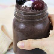 hand holding a mason jar with a cherry smoothie topped with cherries, coconut whip and purple straws.