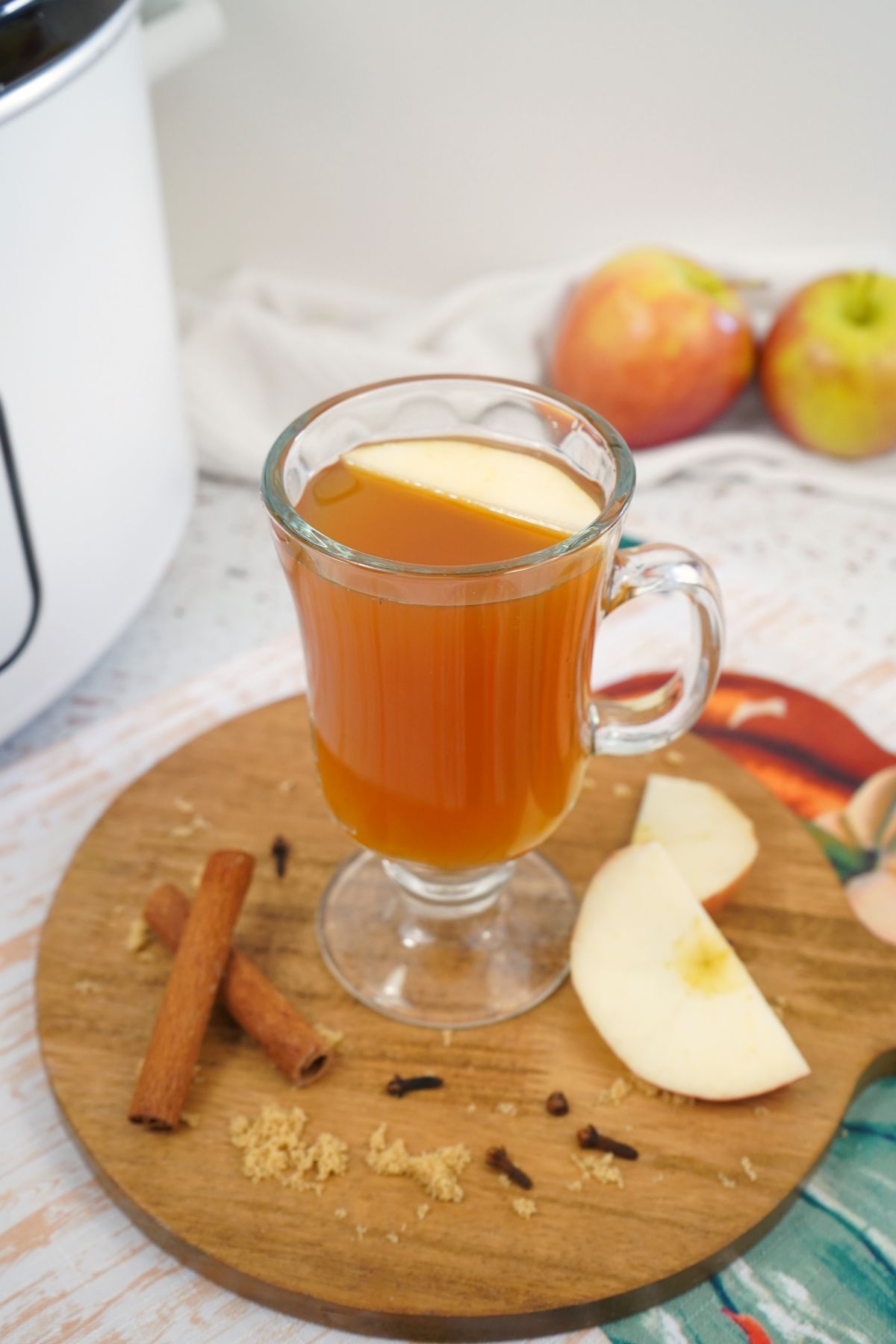 Glass of apple cider on a wooden cutting board with apples and cinnamon sticks around it.