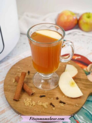 Glass of apple cider on a wooden cutting board with apples and cinnamon sticks around it.