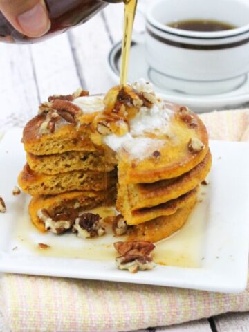 pumpkin pancakes stacked on a white plate with syrup being poured over them