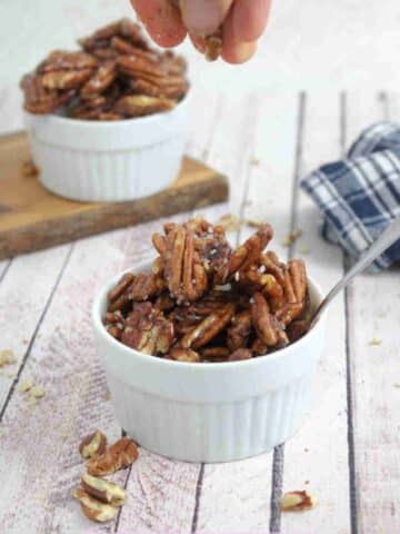 White ramekin with maple candied pecans and a spoon in it with more pecans and a blue linen behind it.