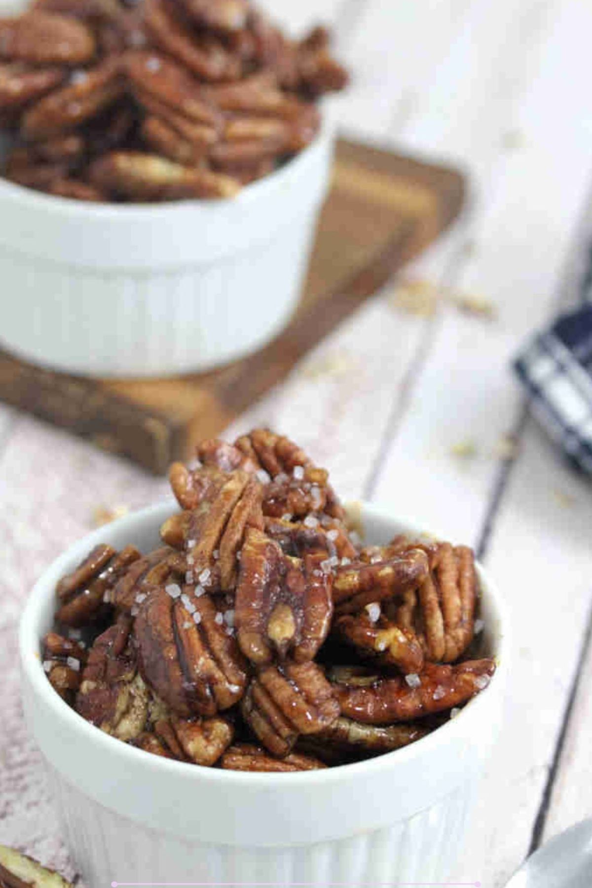 White ramekin with maple candied pecans and a spoon in it with more pecans and a blue linen behind it.