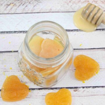 Three honey lemon cough drops around a jar of more cough drops with a honey stick, fresh lemon and a blue linen behind them.