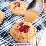 Cooling rack on a blue linen with multiple peanut butter thumbprint cookies topped with jam and a spoon of peanut butter above the first