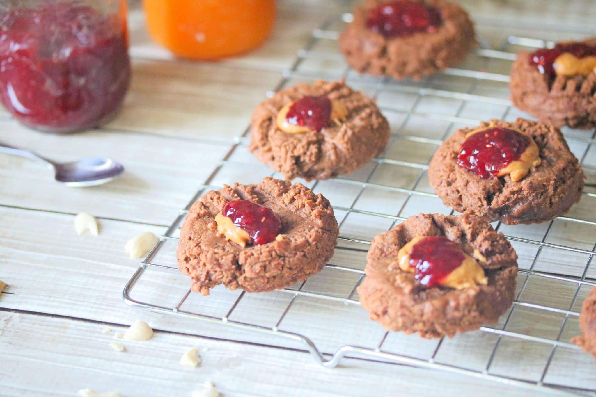 Cooling rack with flourless peanut butter thumbprint cookies topped with jam with a jar of jam and a spoon behind them.