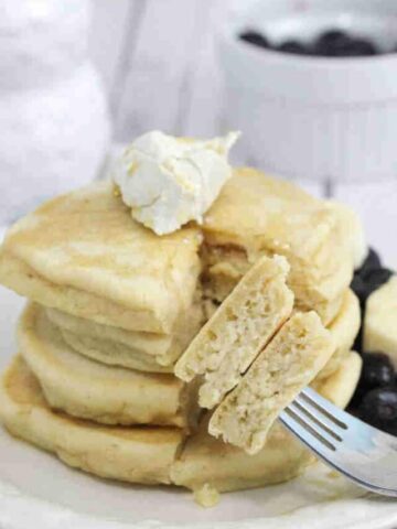 Fluffy pancakes on a white plate with a piece cut out on a fork and maple syrup and butter on the pancakes.