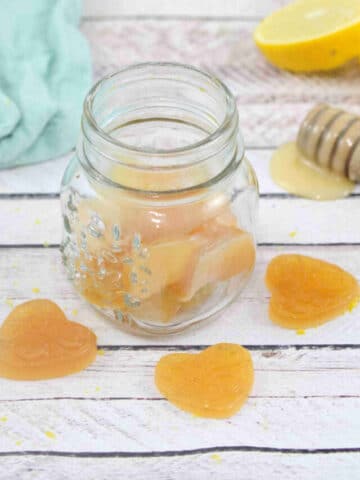 Pin image with text: three honey lemon cough drops around a jar of more cough drops with a honey stick, fresh lemon and a blue linen behind them