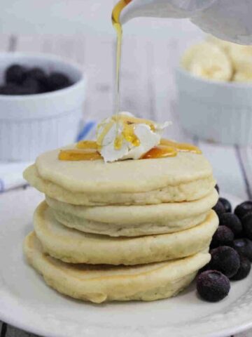 Maple syrup being poured onto fluffy gluten-free pancakes on a white plate topped with butter.